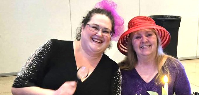 Two Red Hatters in red and purple hats smile side by side at a dinner table.