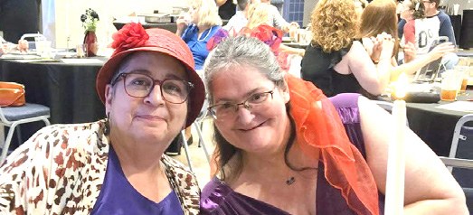 Two Red Hatters, one in a purple hat and one in a red fascinator with a veil, smile for the camera.